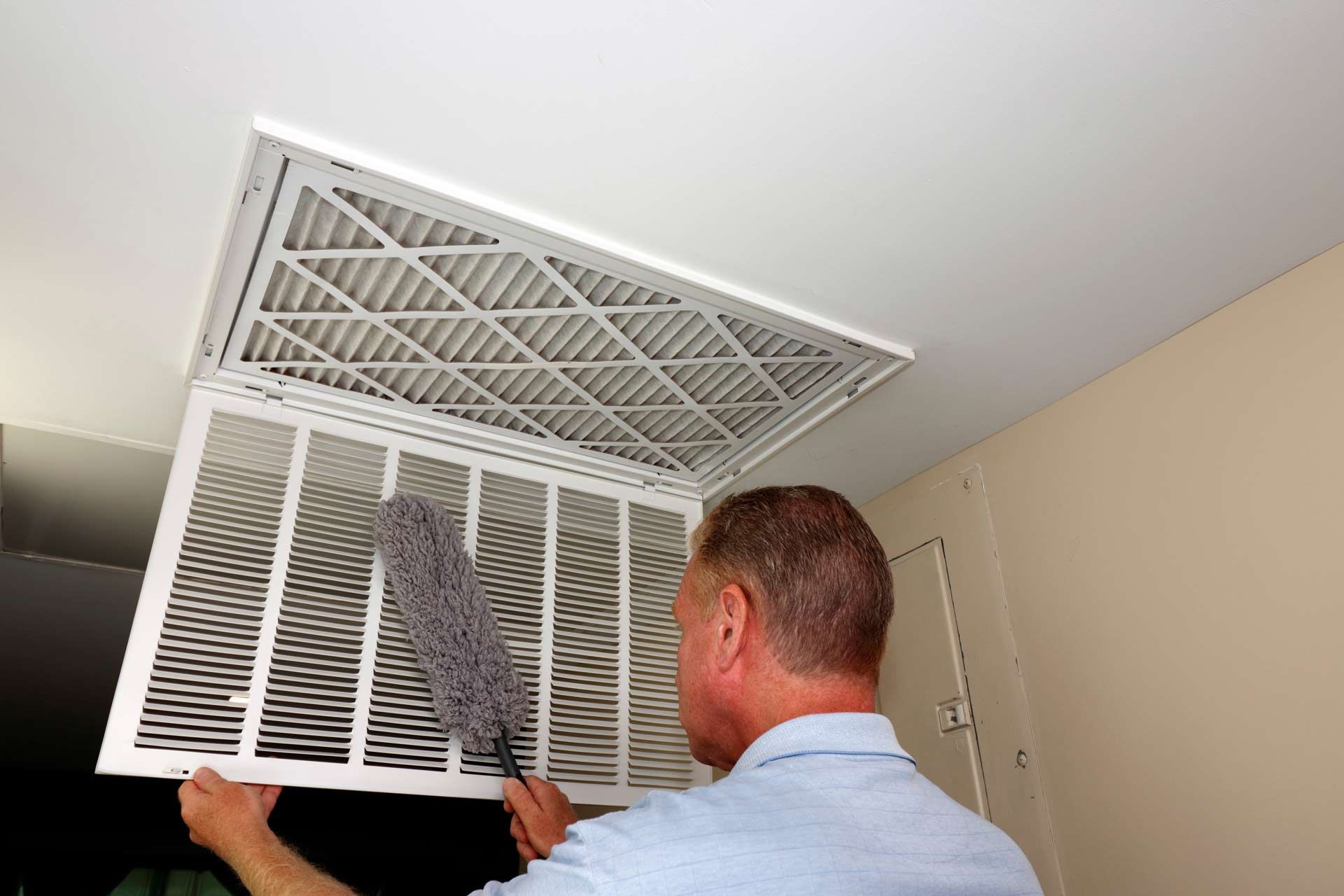 Man cleaning an air filter in a ceiling vent with a duster.