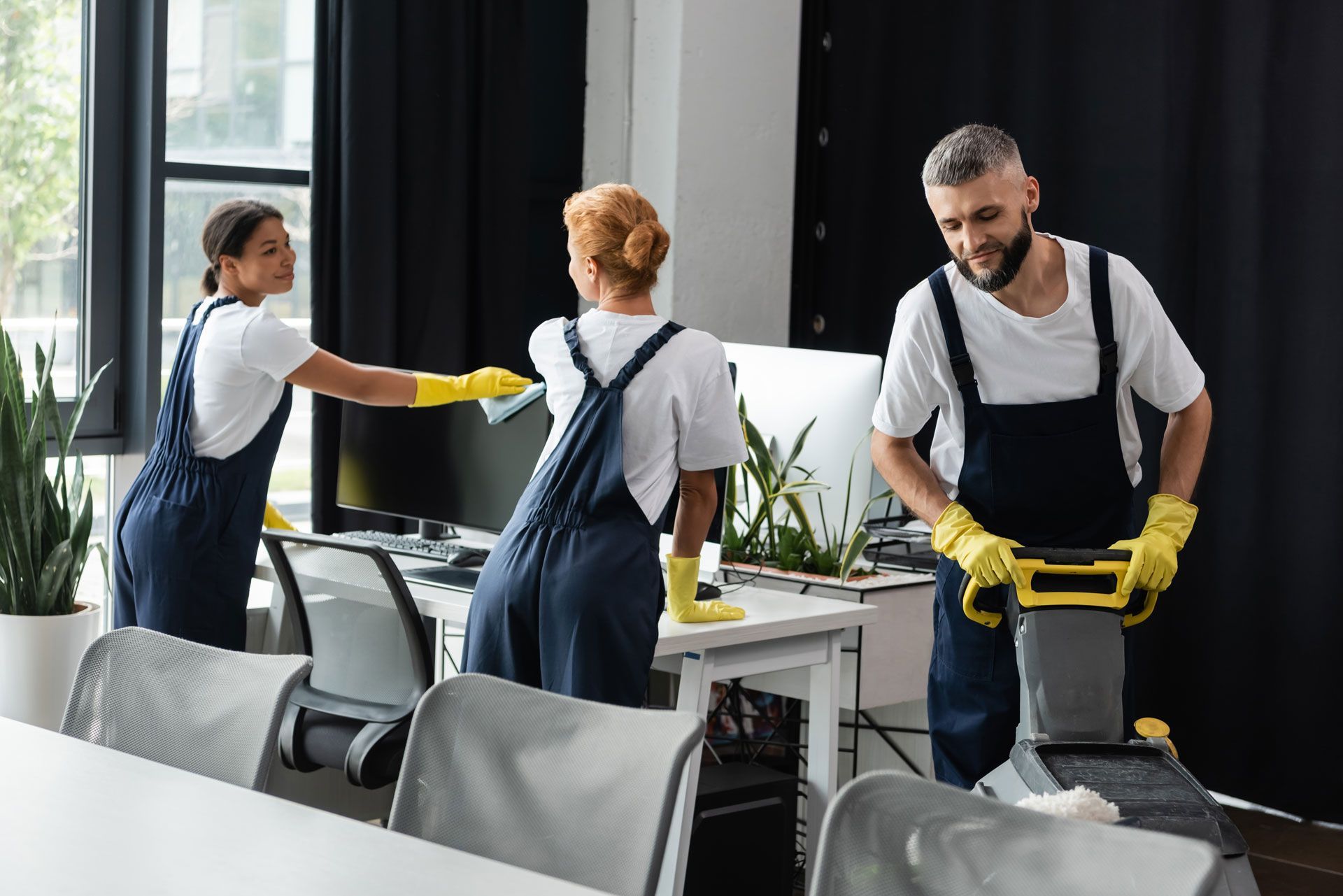 Three people in coveralls cleaning an office: one wipes a desk, one cleans a chair, and one uses a floor machine.