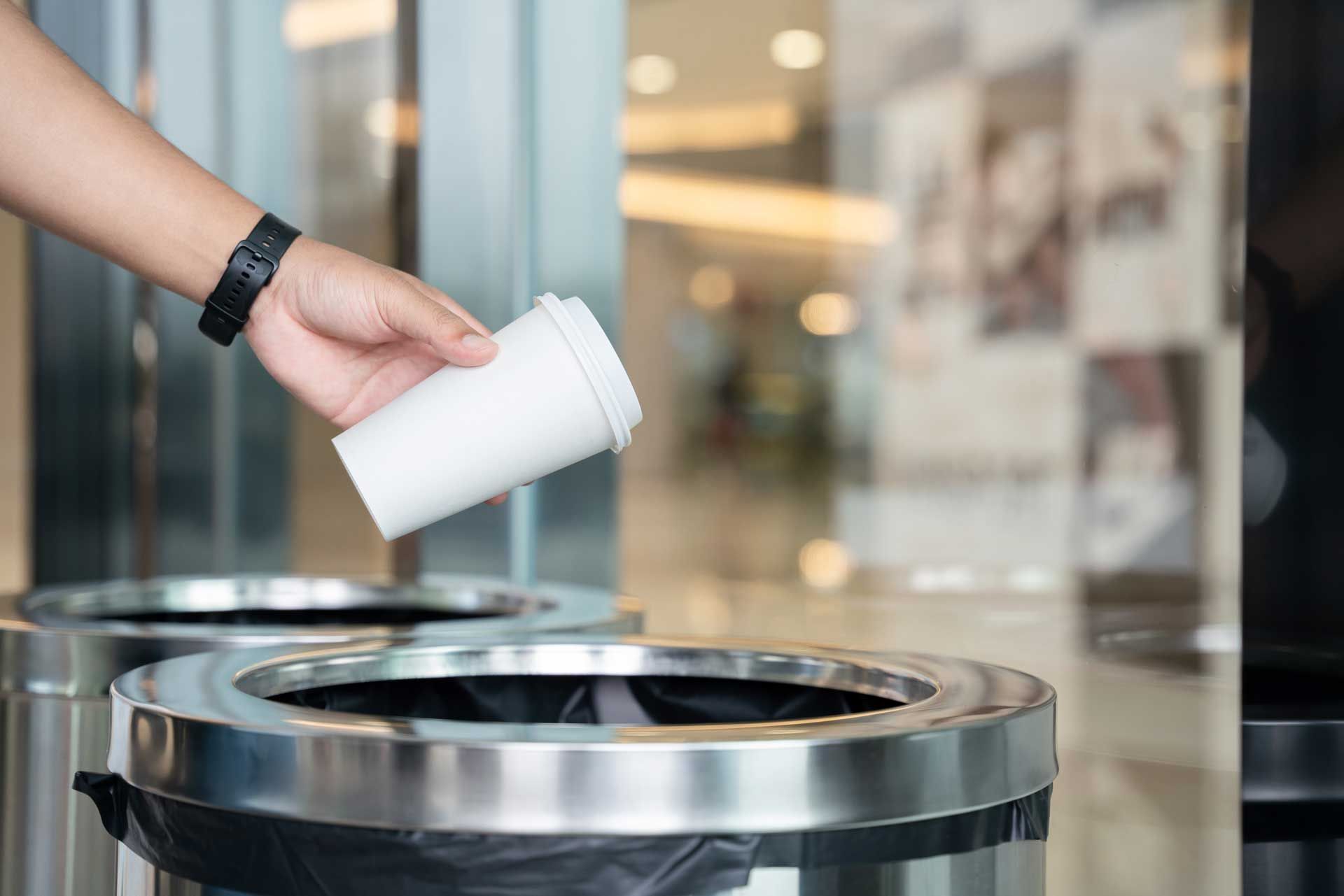 Person throwing a white cup into a metal trash can in a public space.