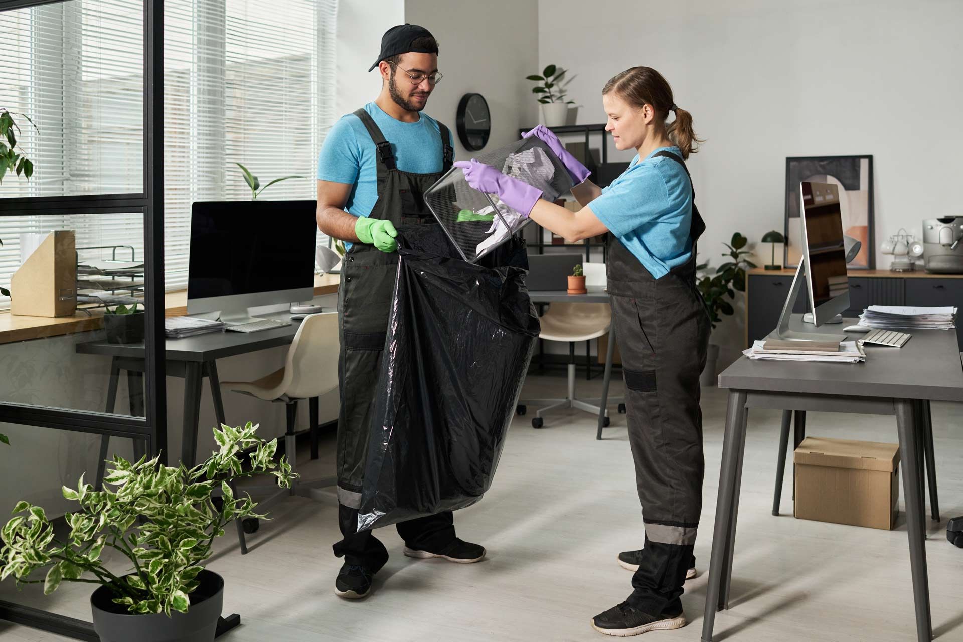 Two people wearing overalls and gloves emptying trash bag in an office.