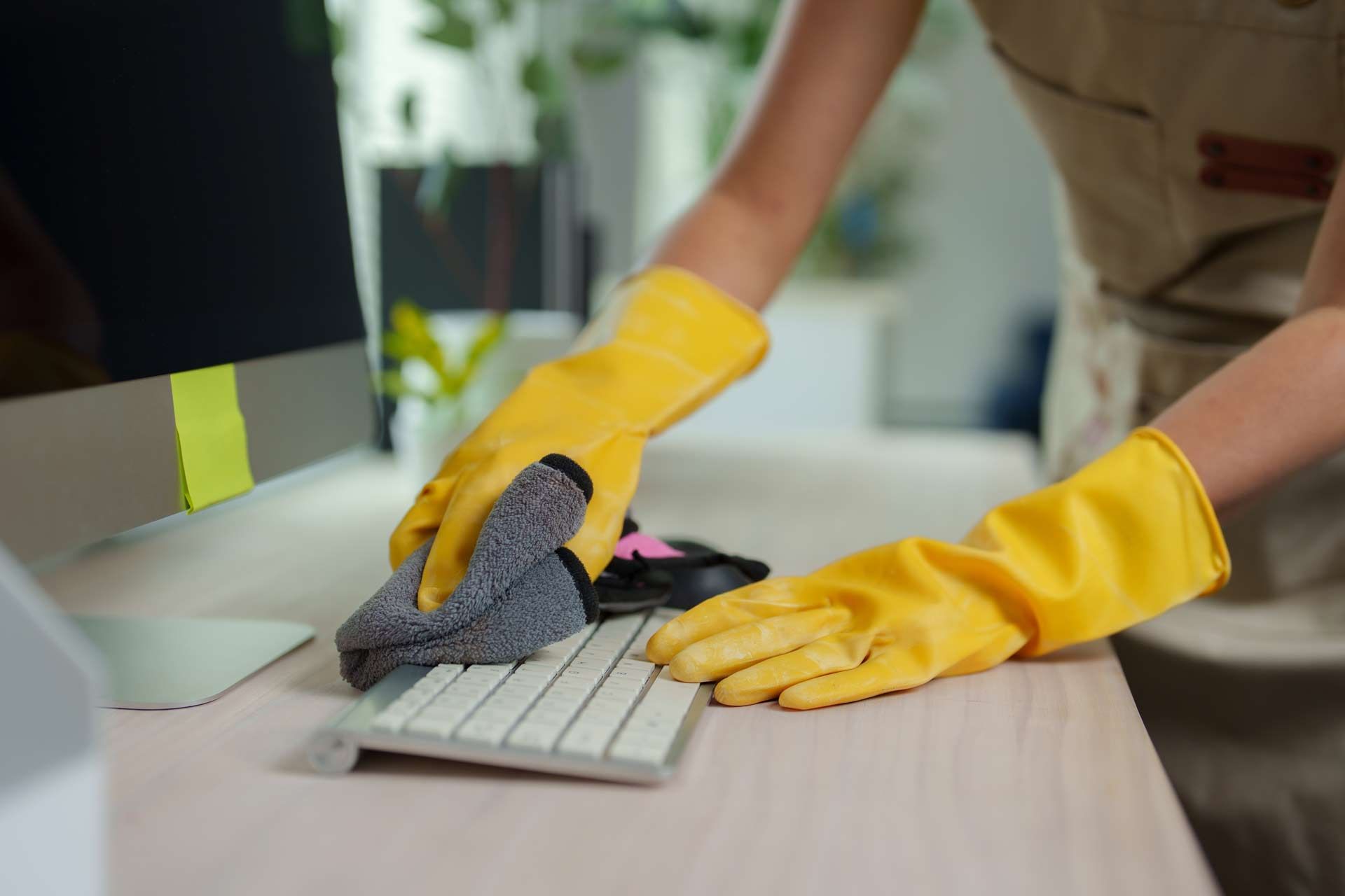 Hands in yellow gloves cleaning a white computer keyboard and desk with a gray cloth.
