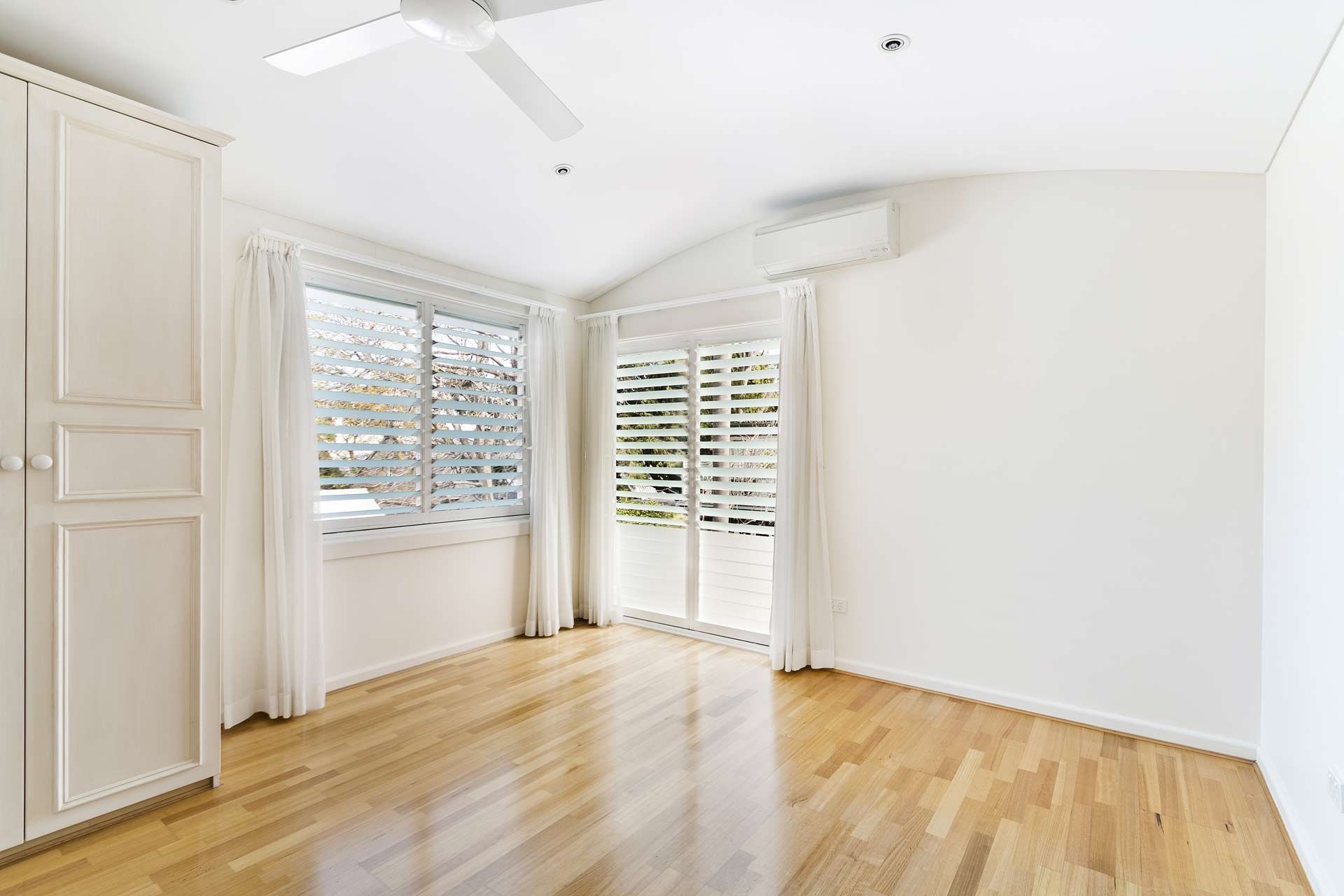 Empty, bright room with hardwood floor, white walls, and closet. Windows with curtains and blinds.