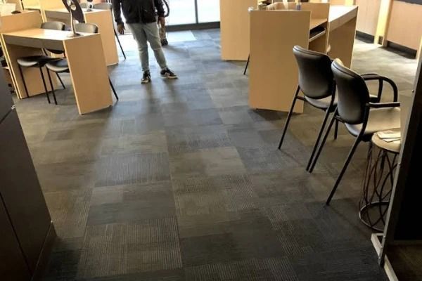 A person stands in a carpeted office with desks, chairs, and light-colored pedestals.