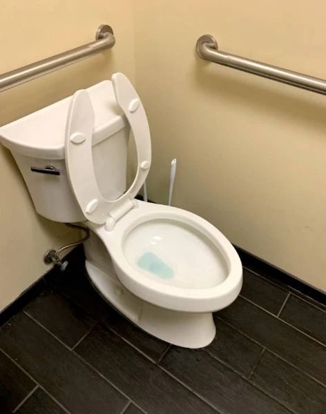 Toilet in a corner with handrails on adjacent walls. The toilet seat is up. Black tile floor.