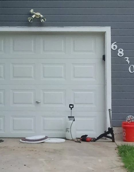 White garage door with tools, a sprayer, and a bucket in front; blue siding and house numbers visible.