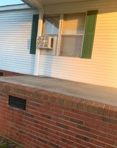 Brick and concrete porch of a house with white siding, green shutters, and a window air conditioner.
