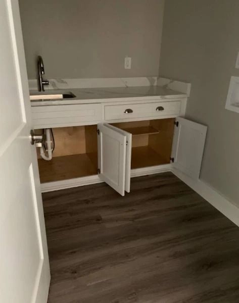 White sink cabinet with opened doors in a laundry room, light countertop, dark wood-look floor, gray walls.