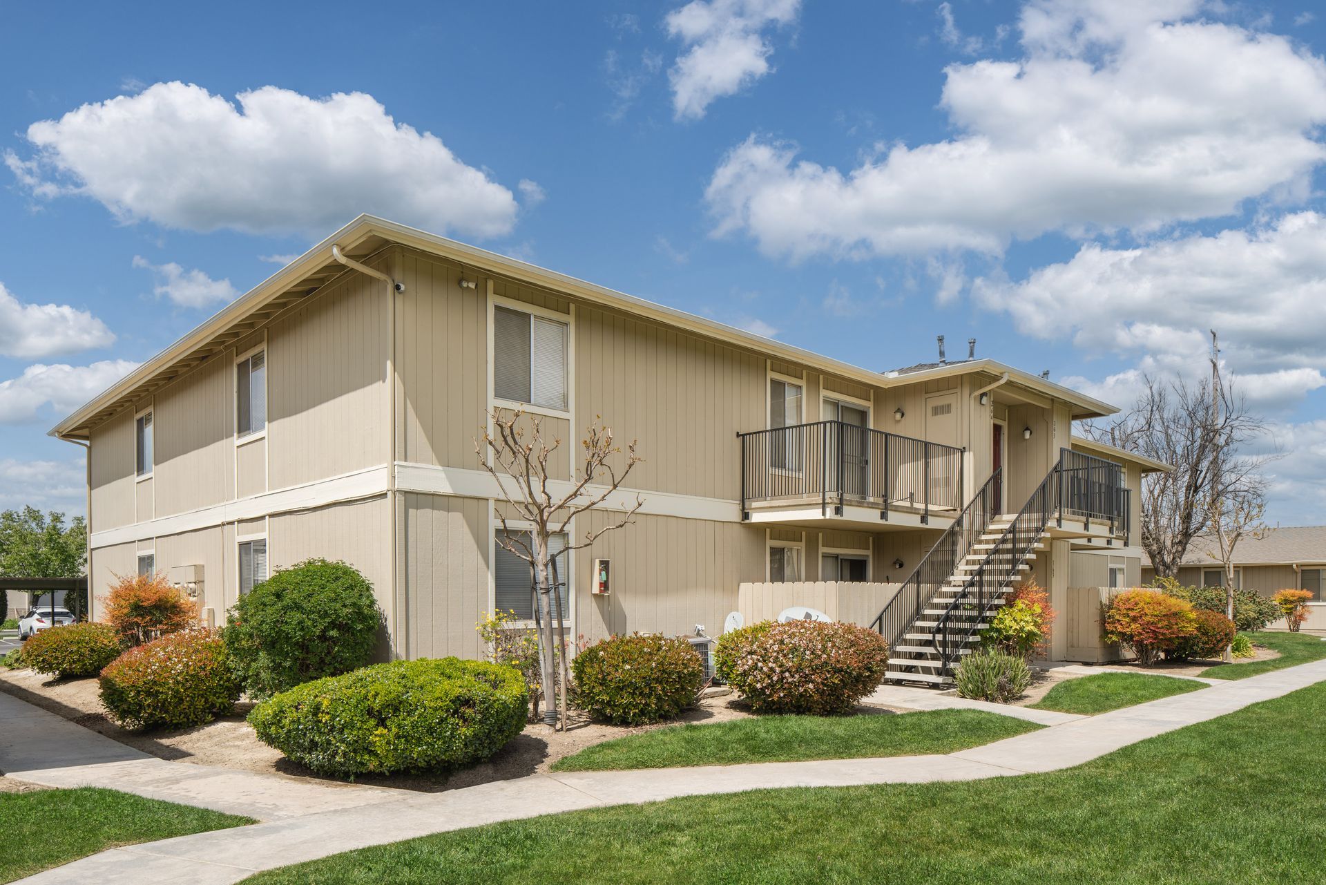 Two-story apartment building with beige siding, staircases, and landscaping under a blue sky with clouds.