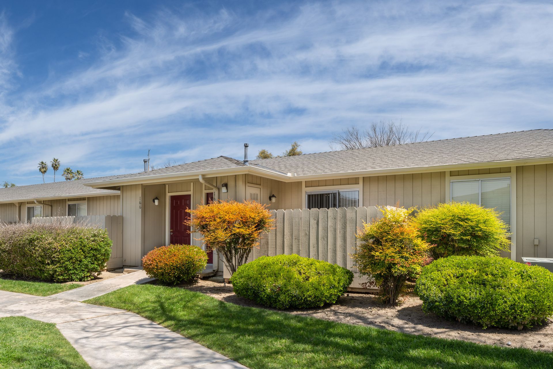 Row of beige townhouses with a red front door, bushes, and a walkway under a blue sky.
