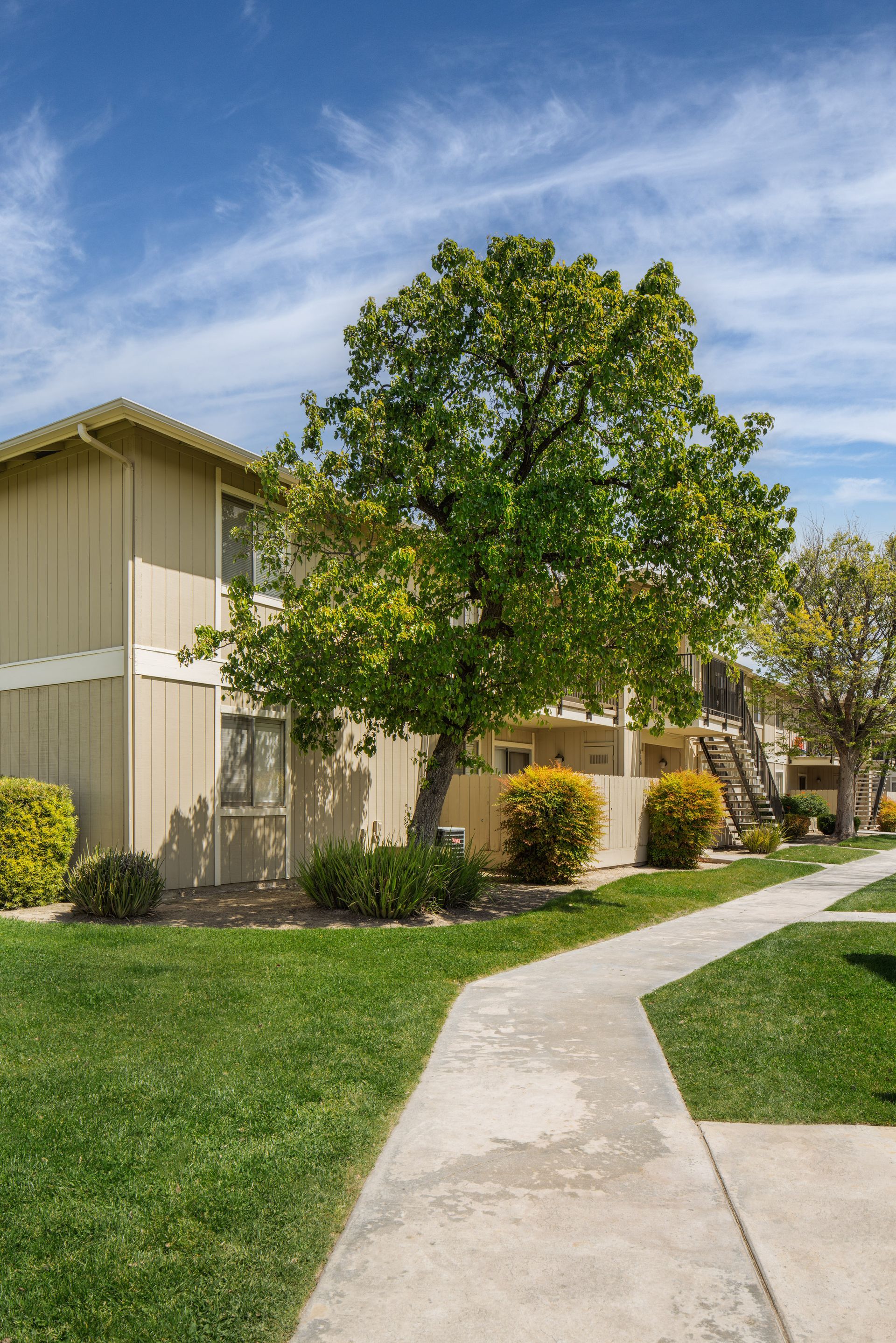 Two-story beige apartment building with a sidewalk and tree, under a blue sky.