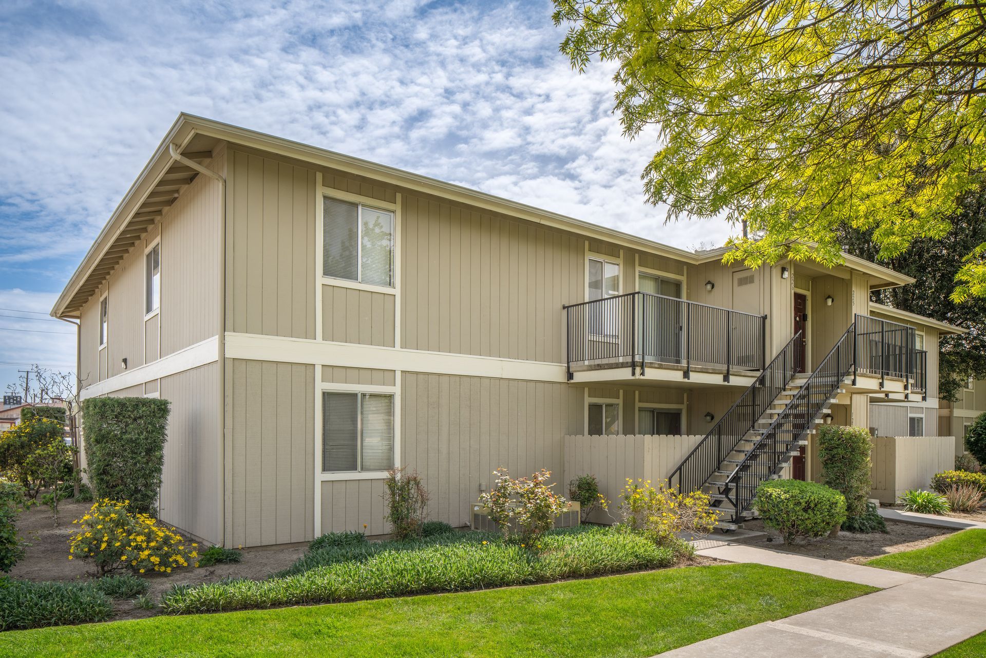 Two-story beige apartment building with balconies, stairs, green lawn, and cloudy sky.
