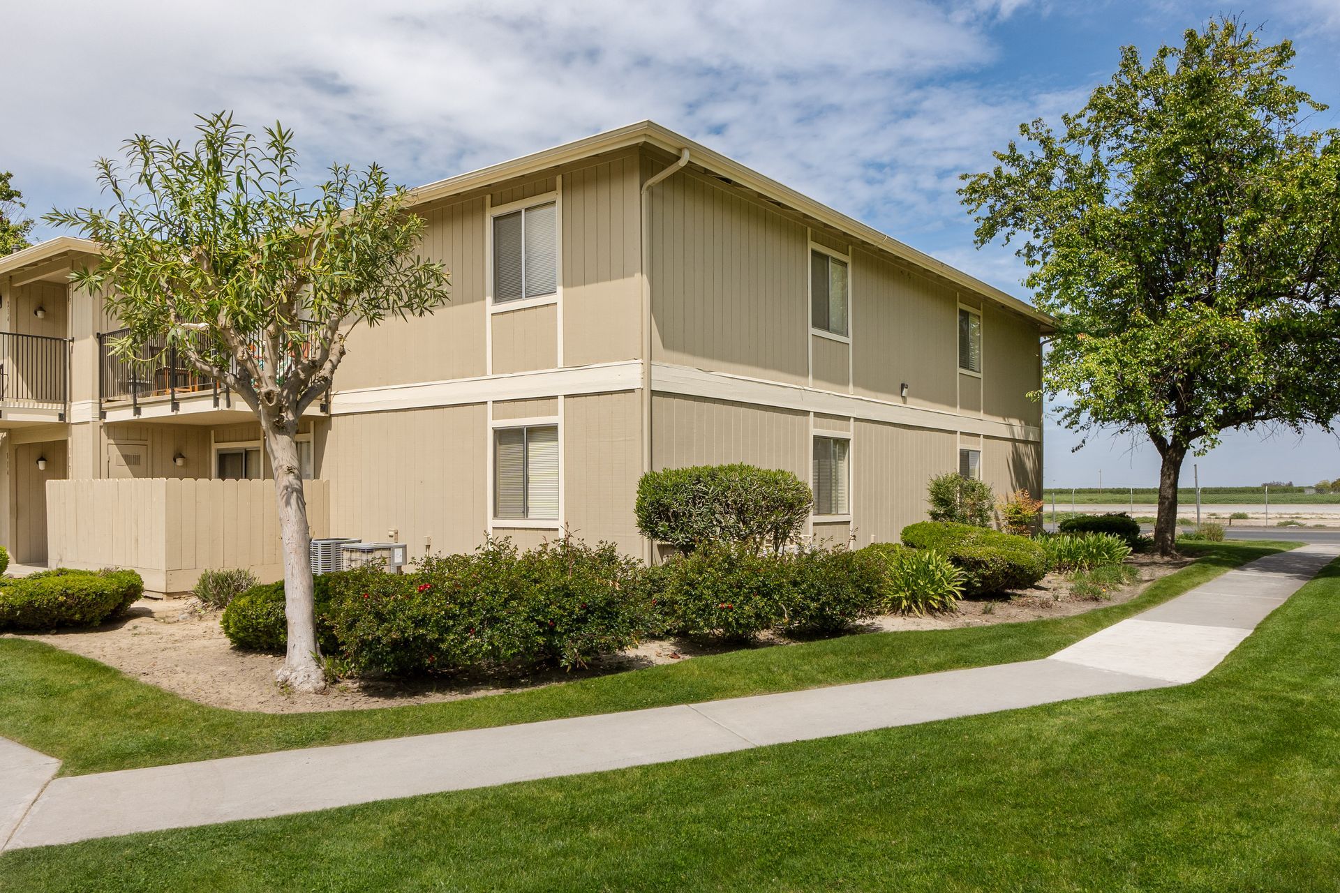 Two-story tan apartment building with windows, green bushes, trees, and a sidewalk on a sunny day.