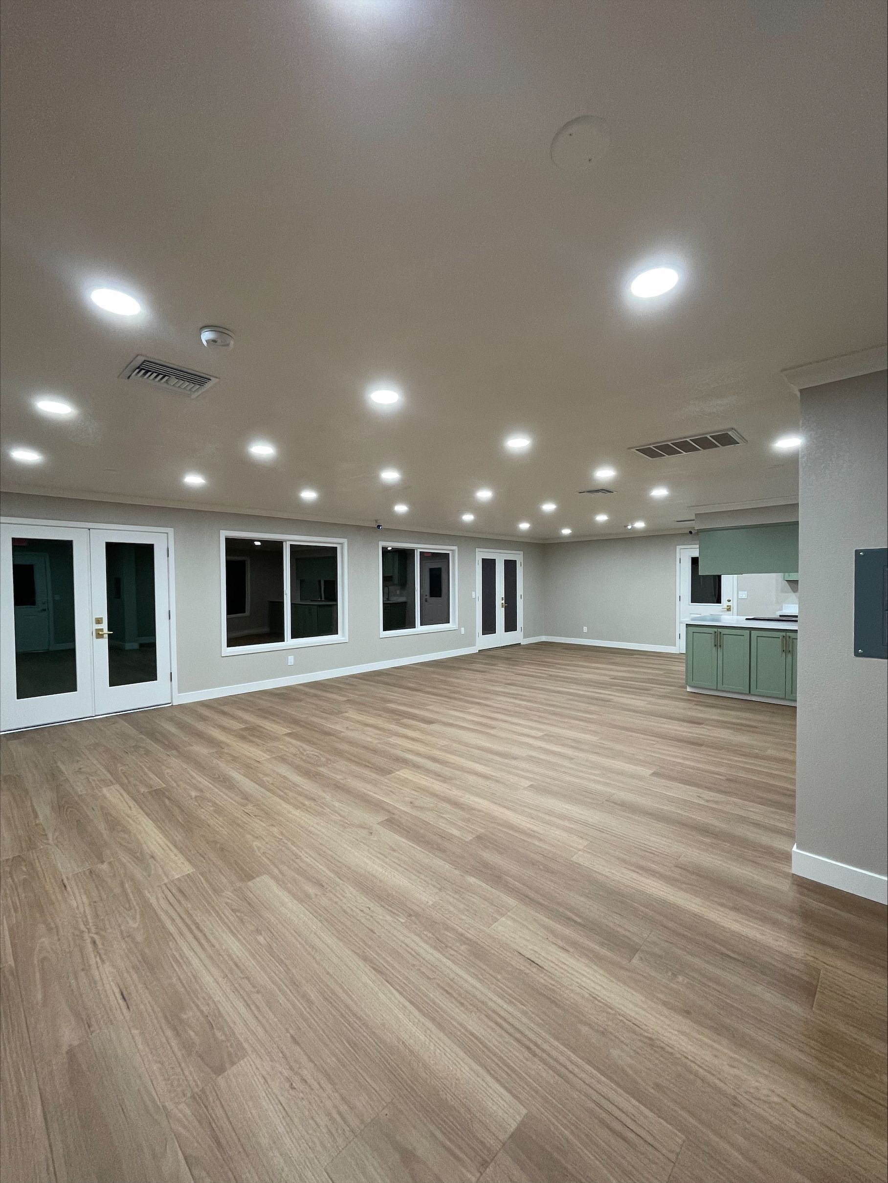 Spacious, empty room with light wood flooring, recessed lighting, and light green accents. French doors and windows.