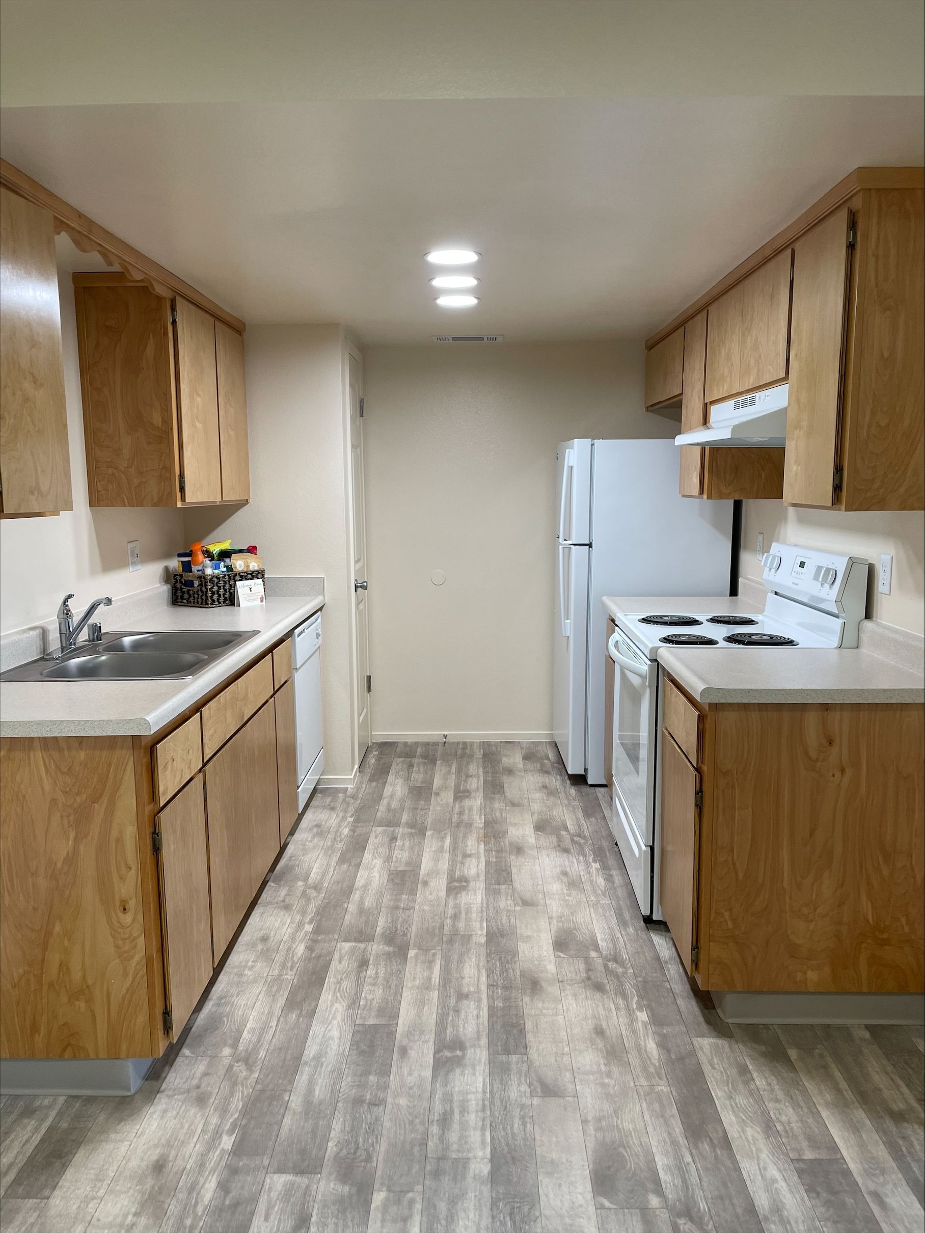 A narrow kitchen with light wood cabinets, white appliances, and gray-toned flooring.