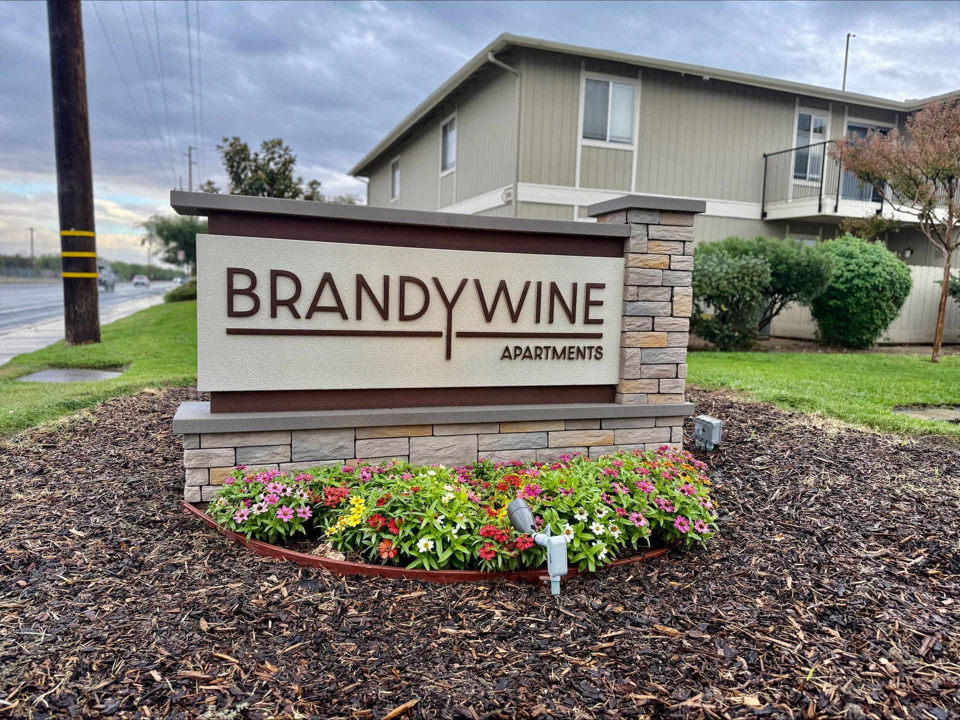 Sign for Brandywine Apartments, featuring stone, flowers, and a two-story apartment building in the background.