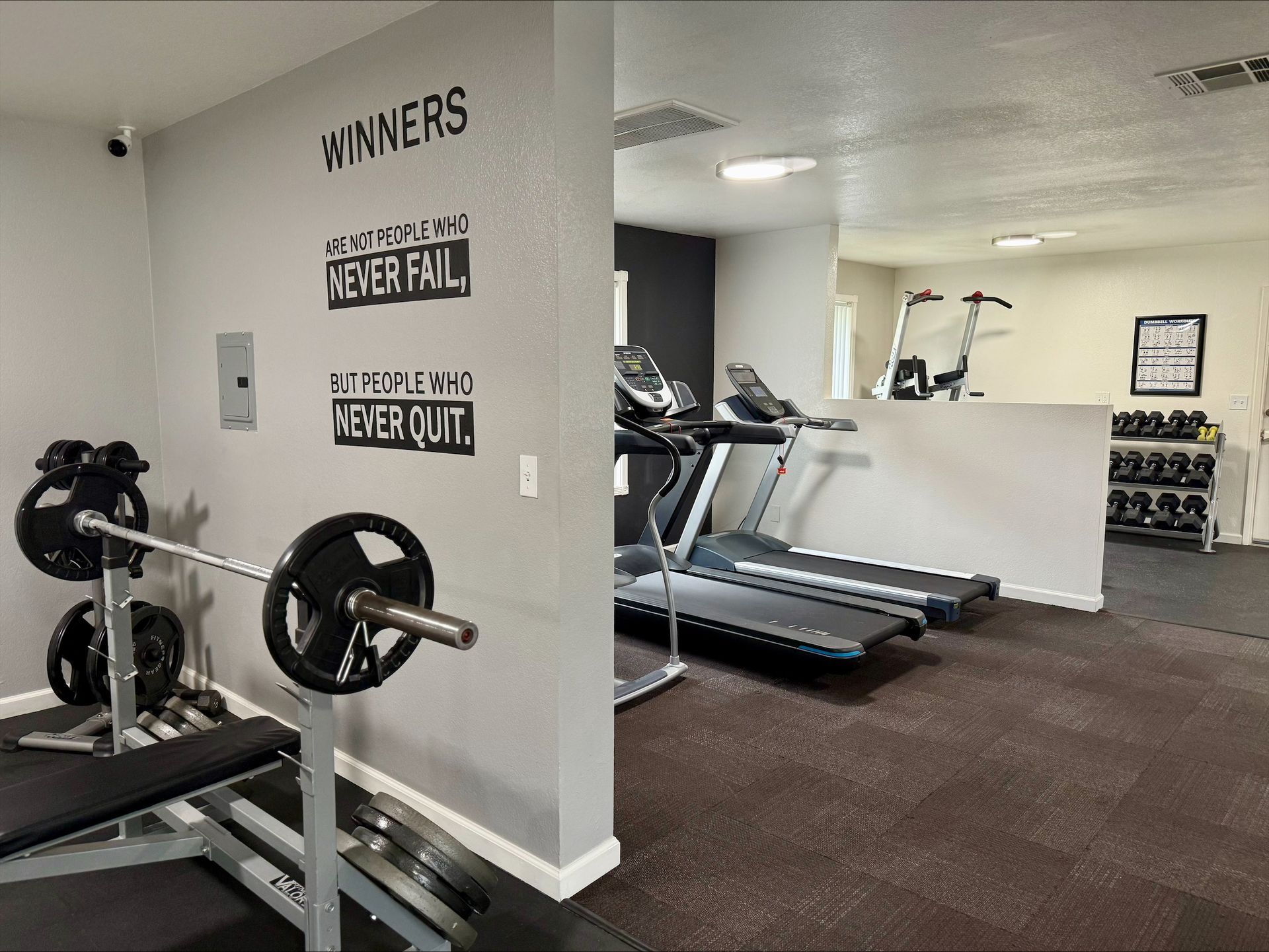 Gym interior with weight bench, treadmill, and dumbbells. Motivational wall art.