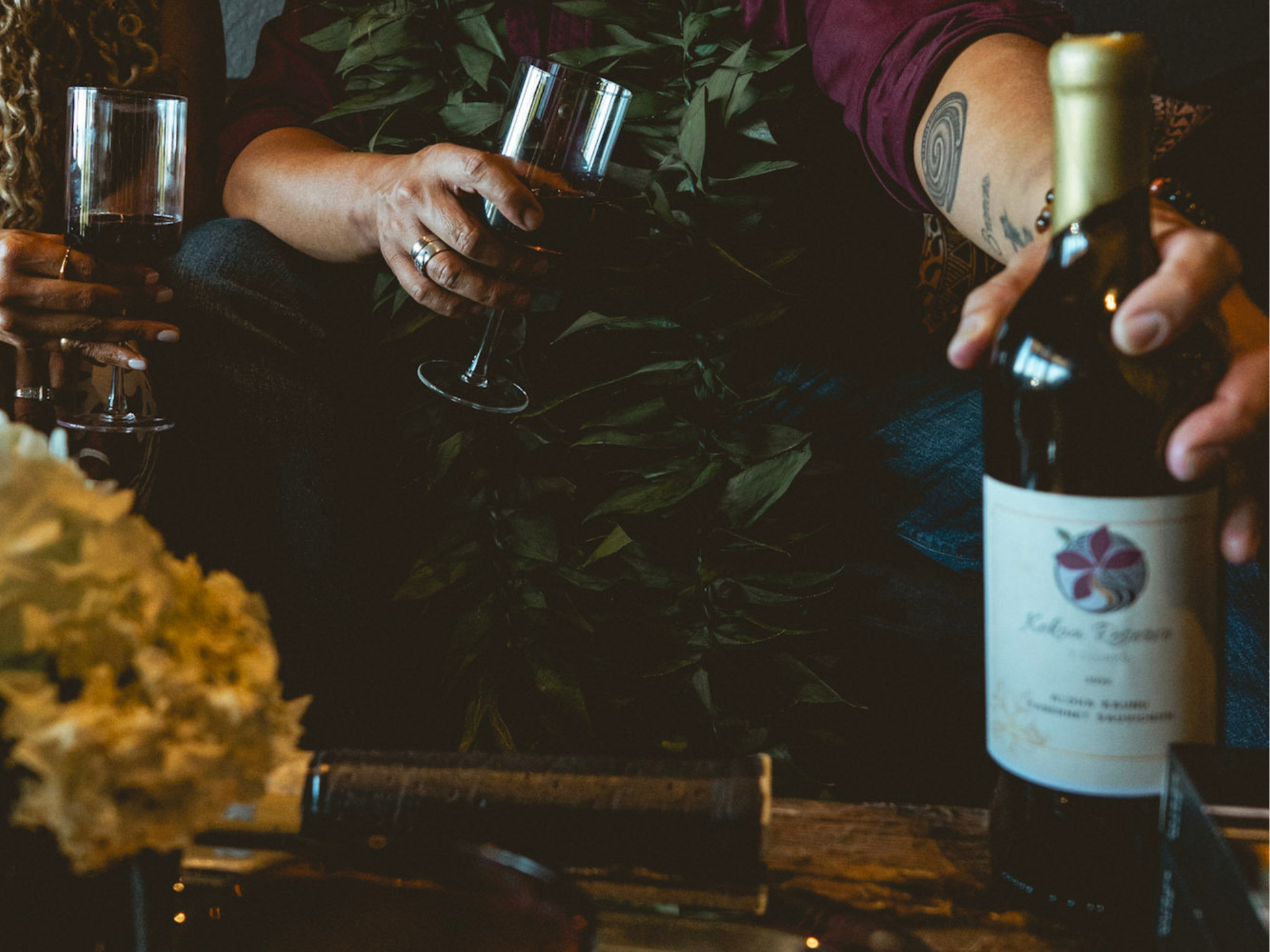 People drinking wine, one holding bottle. Dark setting with foliage.