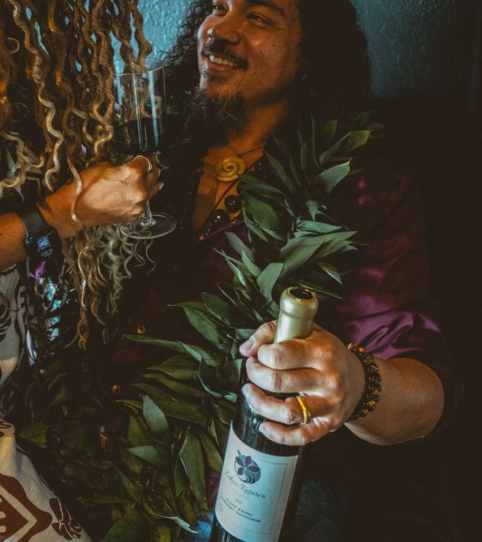 Man holding wine bottle, smiling, wearing leaf lei and bracelets, with someone's hand holding champagne glass.