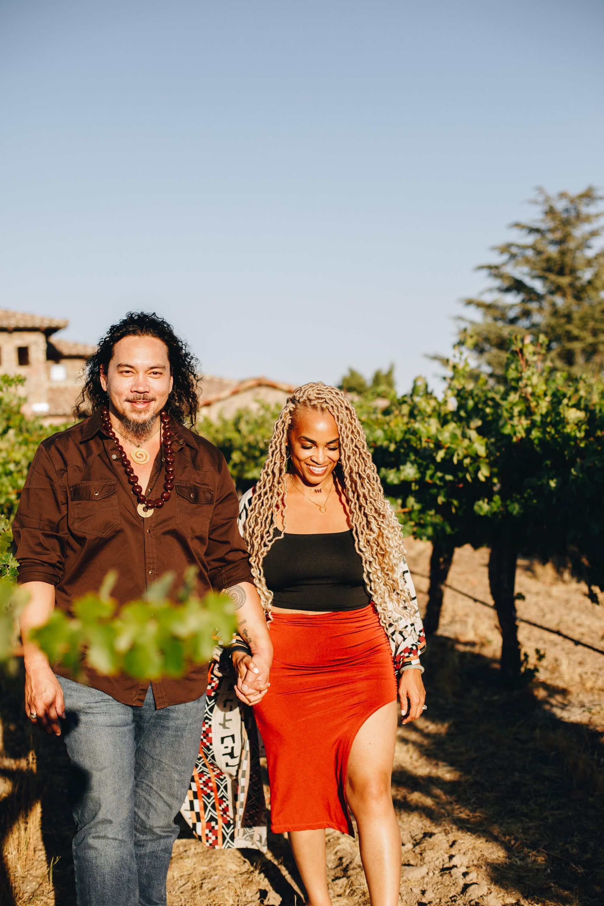 Man holding wine bottle, smiling, wearing leaf lei and bracelets, with someone's hand holding champagne glass.