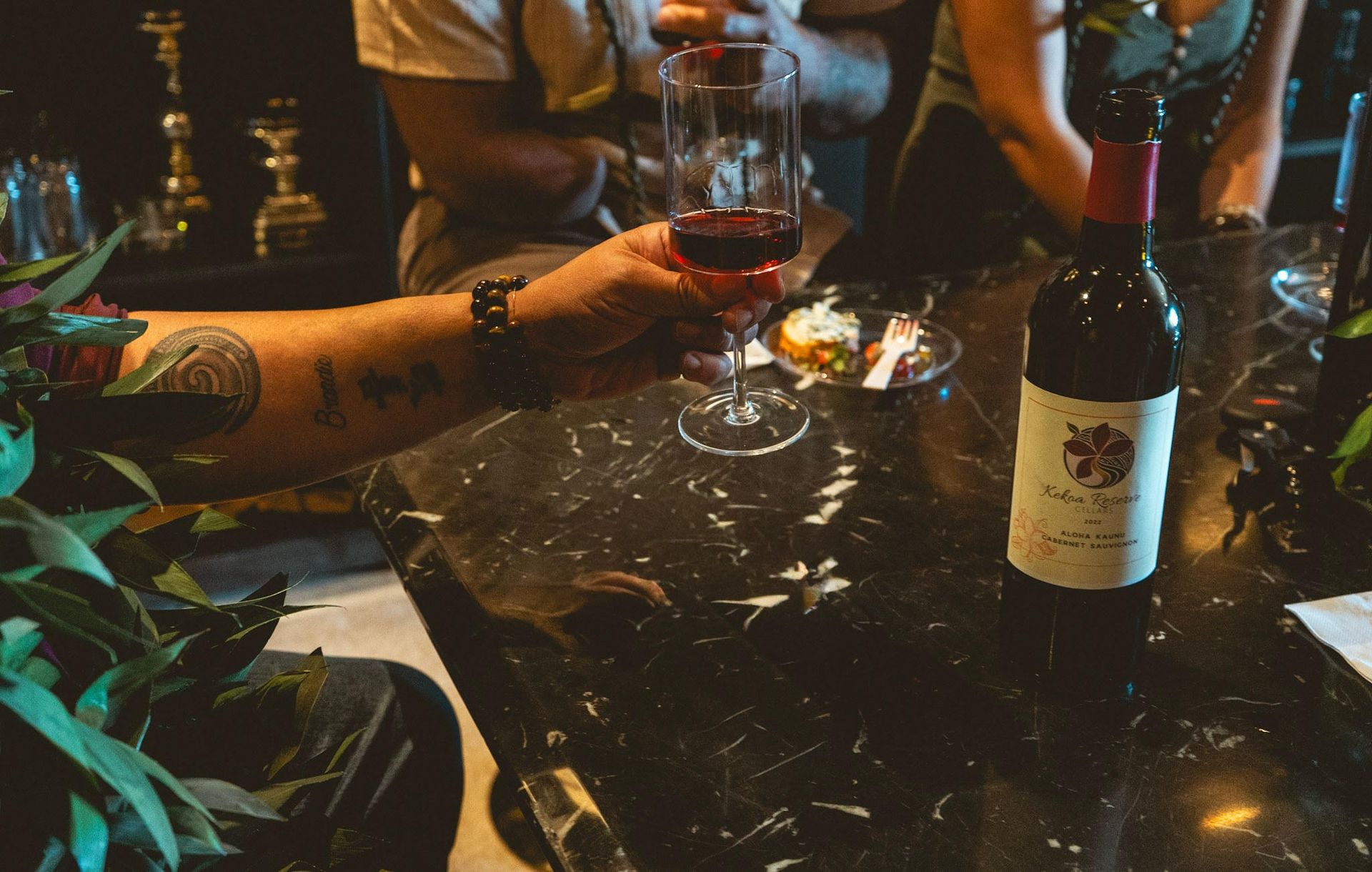 Vineyard leaves in the foreground, with rows of grapevines and a colorful sunset sky in the background.