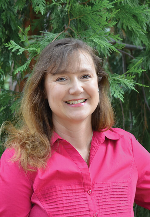 Woman with brown hair smiles, wearing a pink shirt, set against a green leafy background.