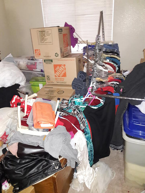 Room with clothes and boxes piled up, including a clothing rack, tan boxes, and various colored garments.