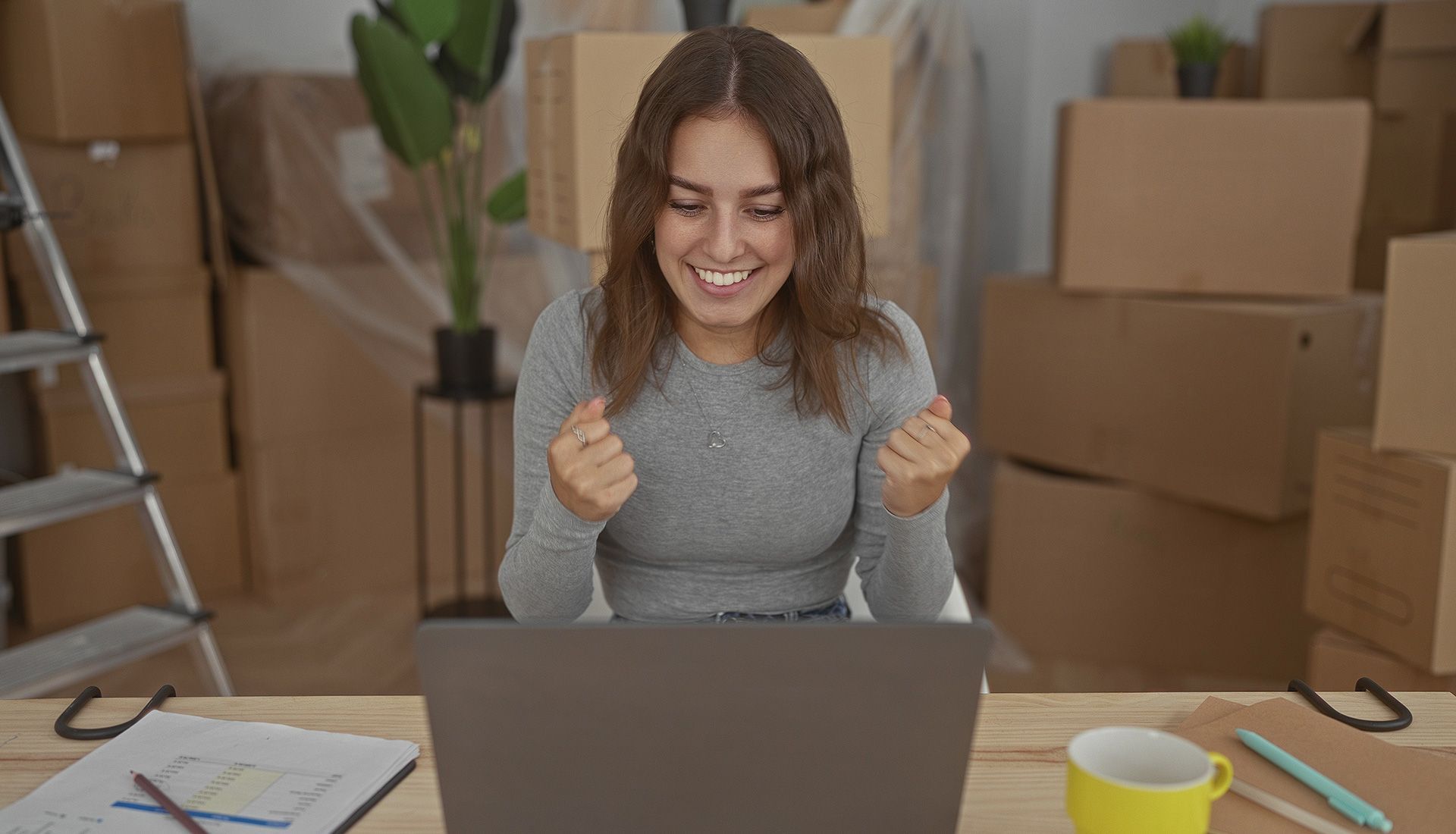 Woman clenching fists, smiling at laptop in room with moving boxes.