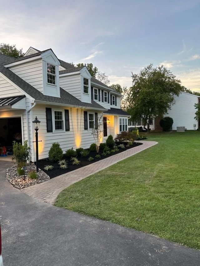 White two-story house with black shutters, lit windows, landscaped front garden, and a curved driveway at dusk