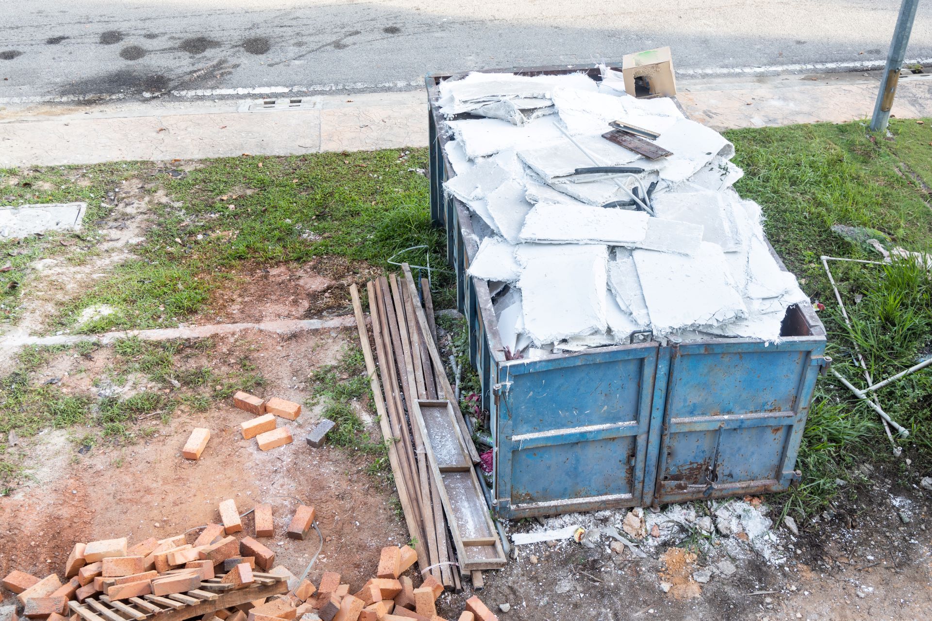A blue dumpster is sitting in the grass next to a pile of bricks.