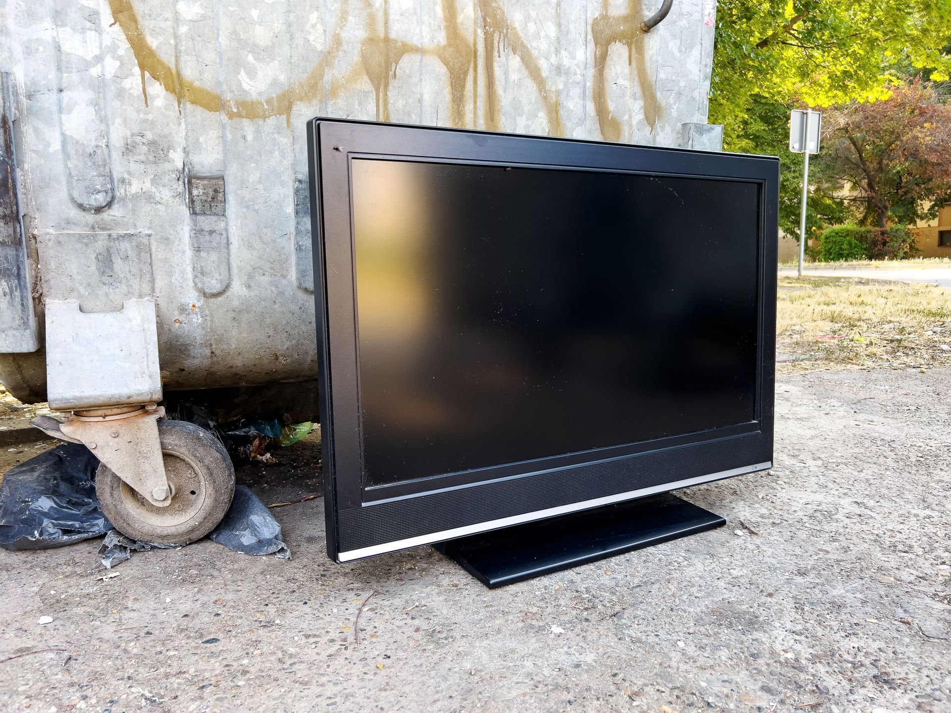 A flat screen tv is sitting on the ground next to a dumpster.