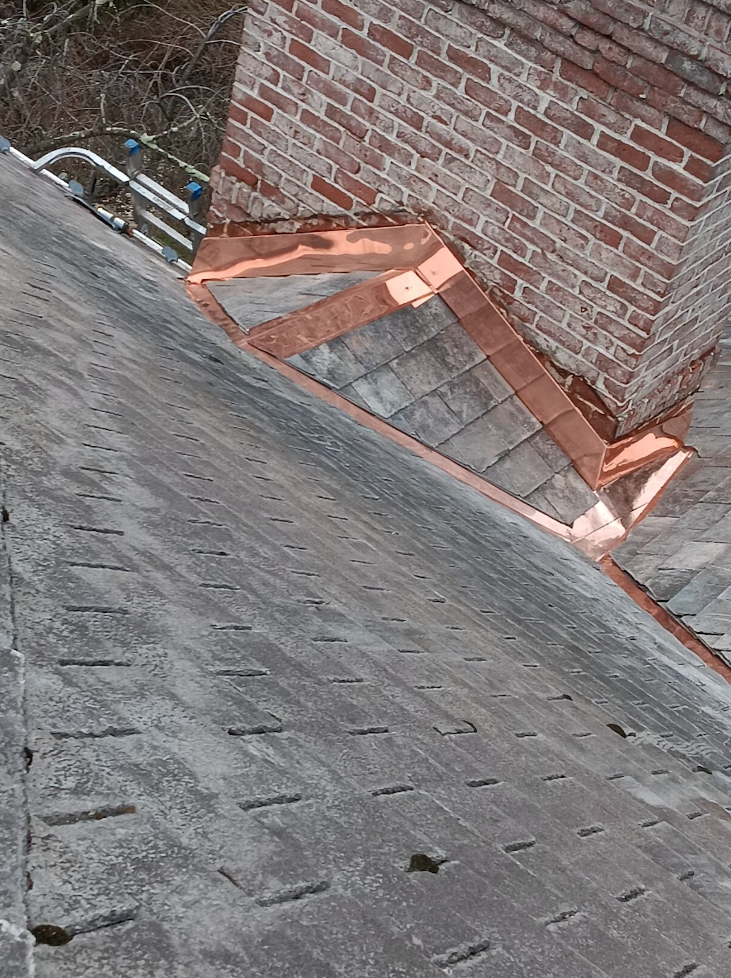 Close-up of an aged gray roof with a copper-lined brick chimney and some other roof-related elements.