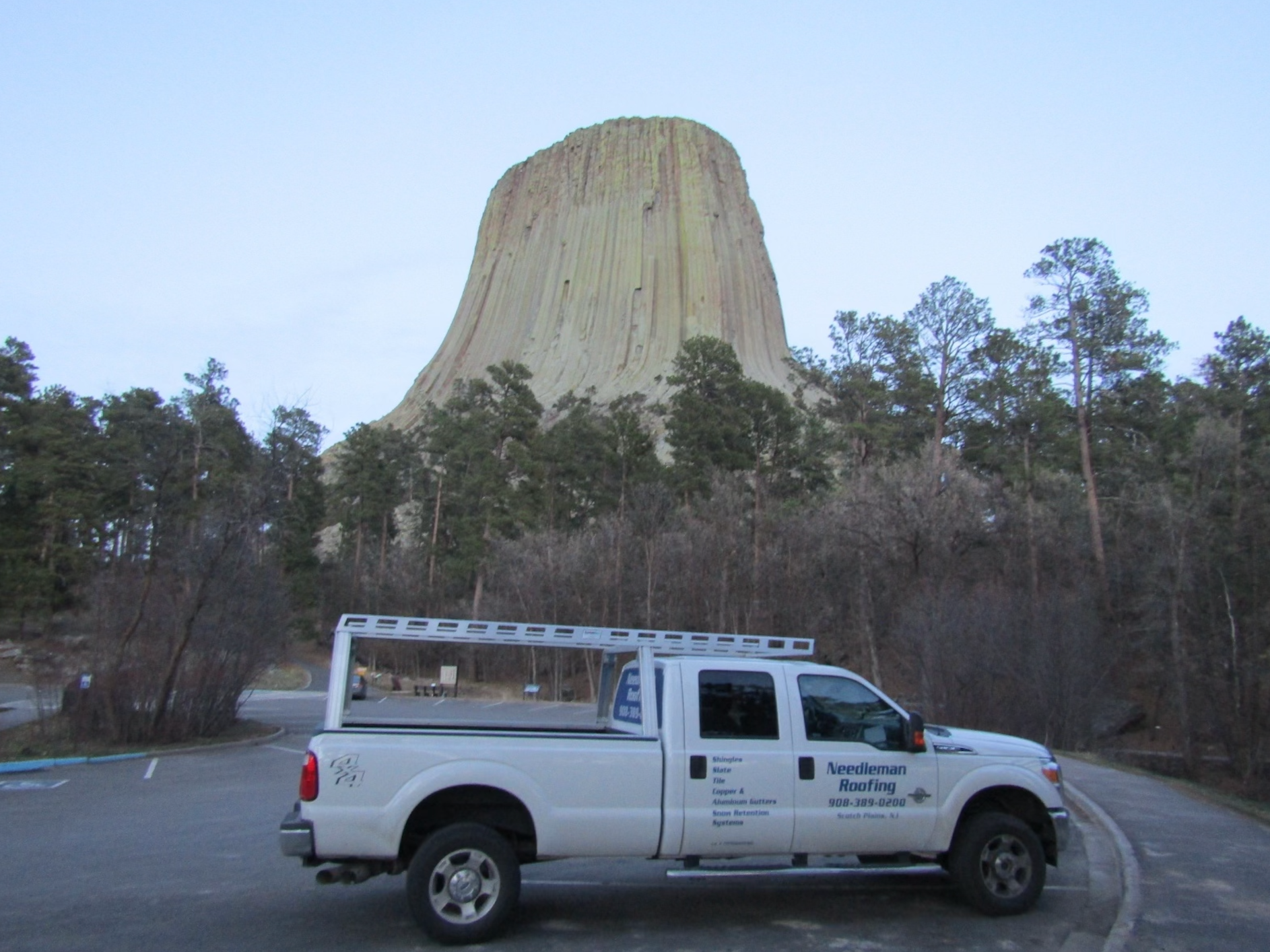 White truck parked with Devils Tower in the background. Cloudy sky, trees.