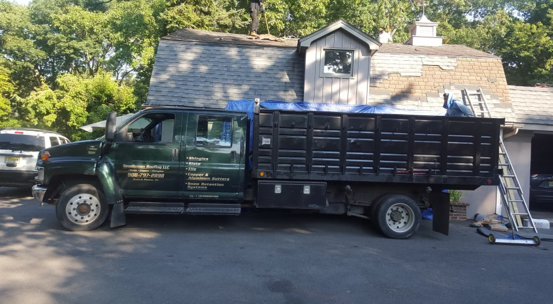 A green dump truck parked near a house with roofing work underway.