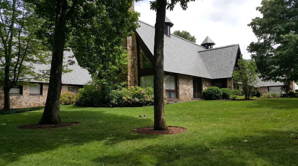 Brick building with a unique roof design, set in a grassy yard with trees.