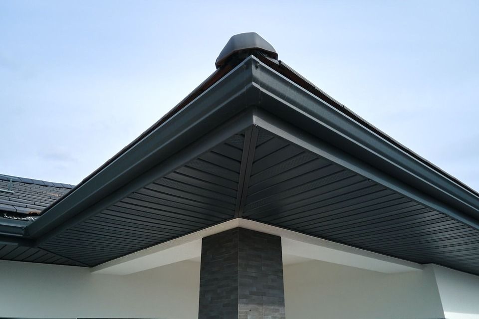Corner of a house with dark gray gutters, soffit, and trim. White wall, black tiled roof, against a cloudy sky.