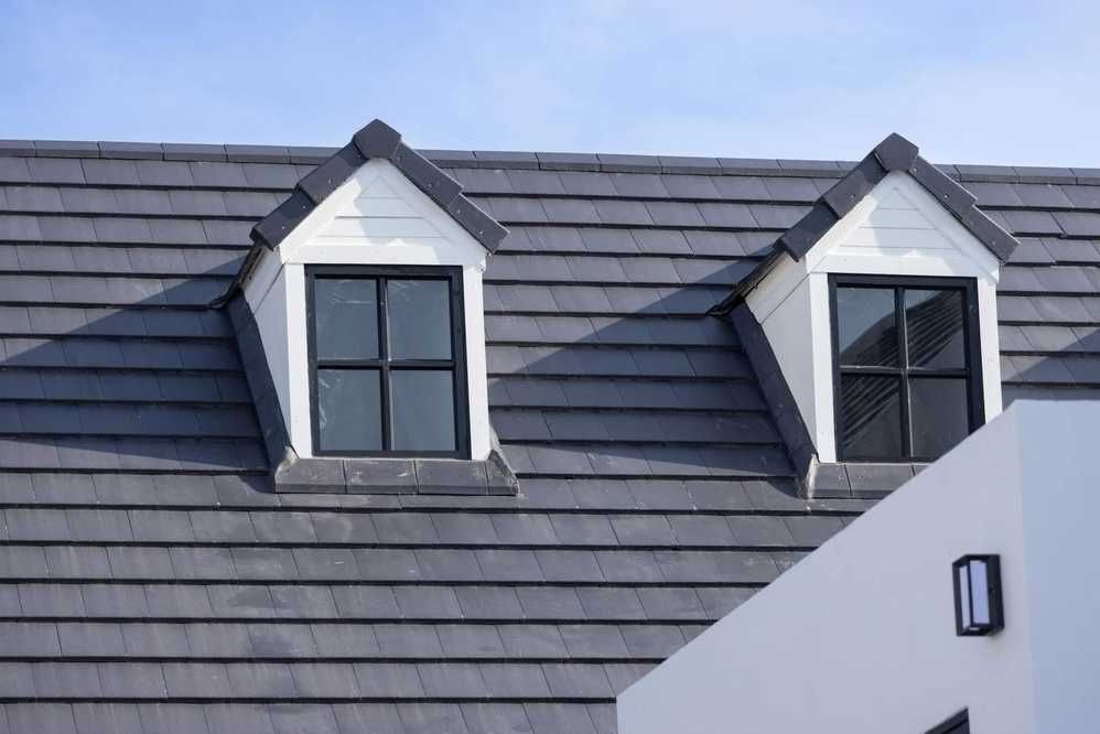 Two white dormer windows with black frames on a gray-tiled roof, with a white wall in the foreground.