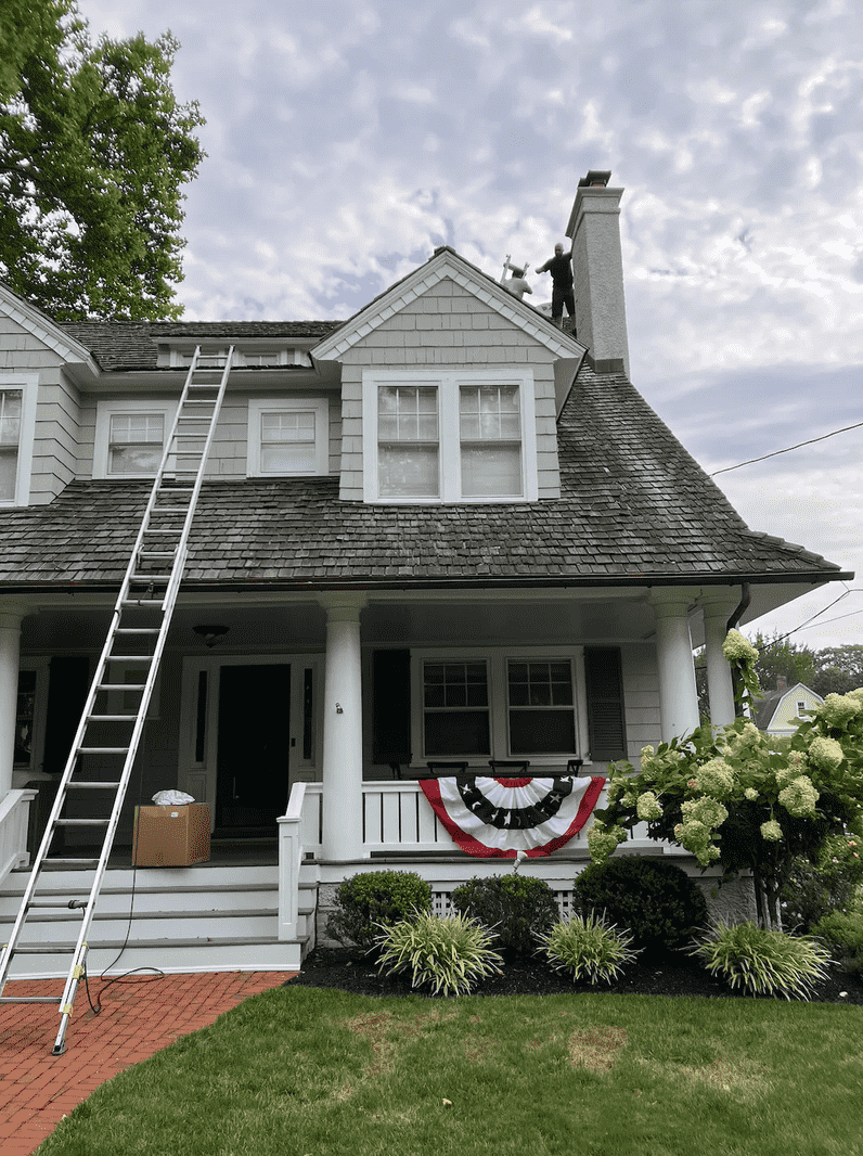 Ladder against a two-story gray house with a person on the roof. American flag bunting hangs on the porch.