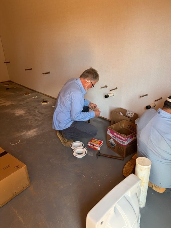 Person kneeling on a floor, sorting tools and supplies beside a bare wall in an unfinished room.