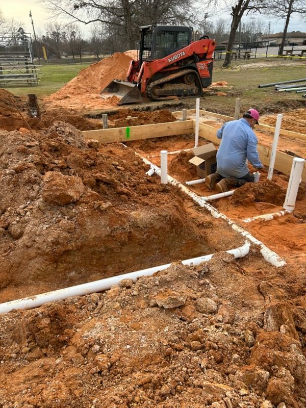 Construction worker installing white PVC pipes in a muddy excavation beside an orange skid steer.