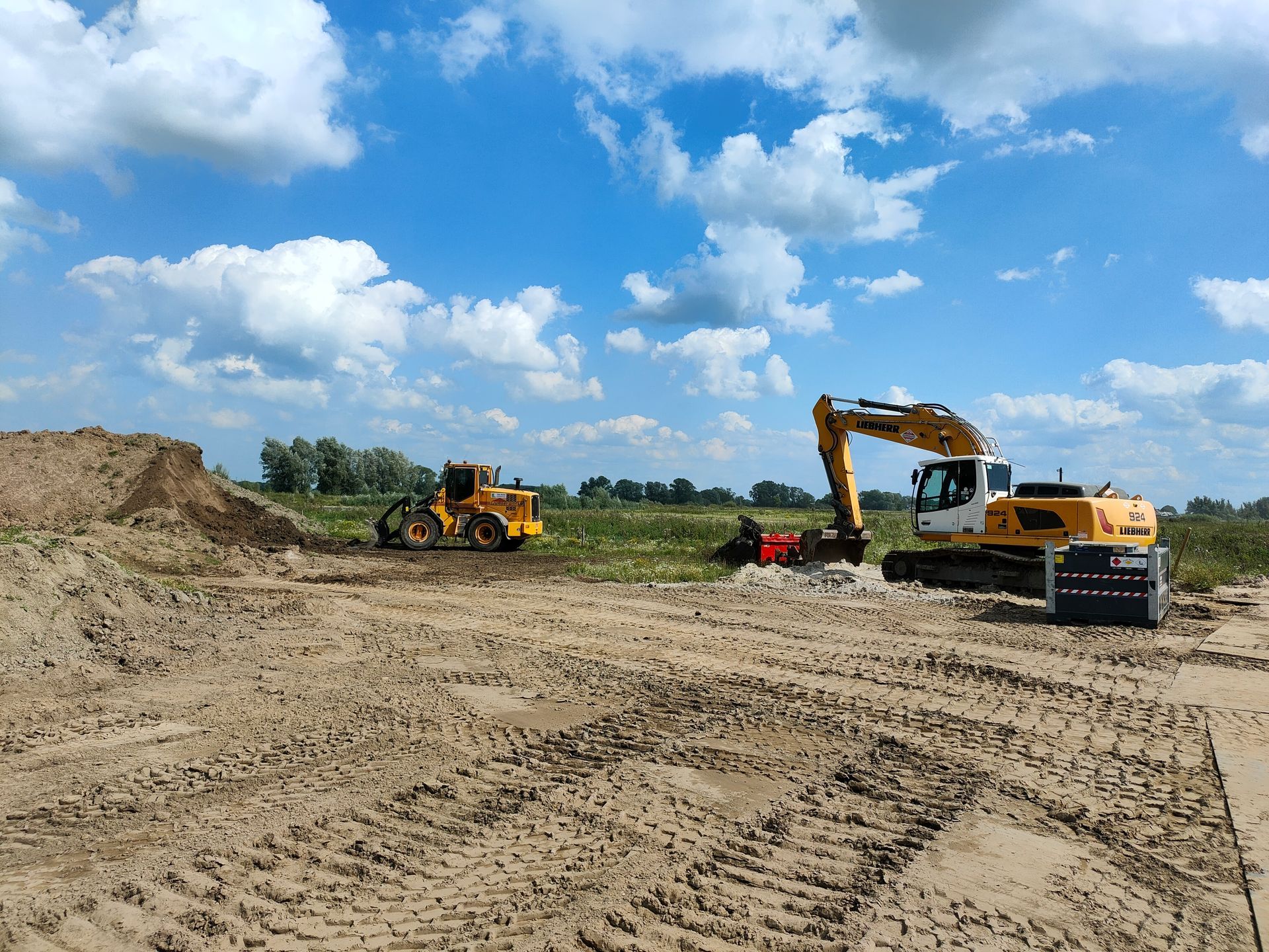 Bouwplaats met graafmachine en wals op zandgrond, blauwe lucht en wolken.