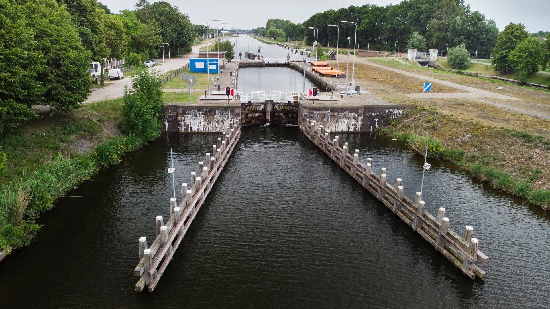 Een sluis met open houten deuren. Het water stroomt erdoorheen, geflankeerd door betonnen muren en groen. Bewolkte lucht.