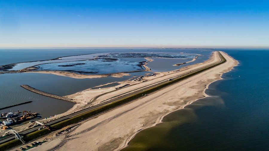 Luchtfoto van een lange, zandige landtong met een weg die de blauwe zee in loopt.