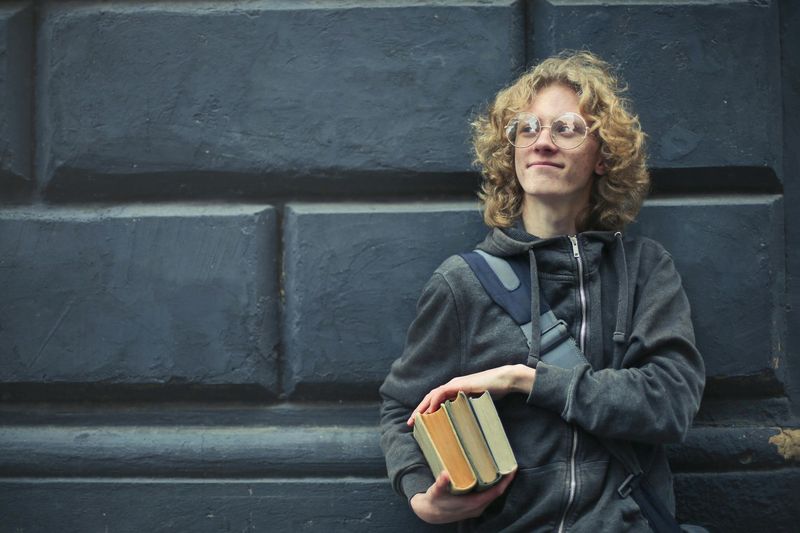 Person with curly hair holding books, leaning against a dark wall and looking upward.