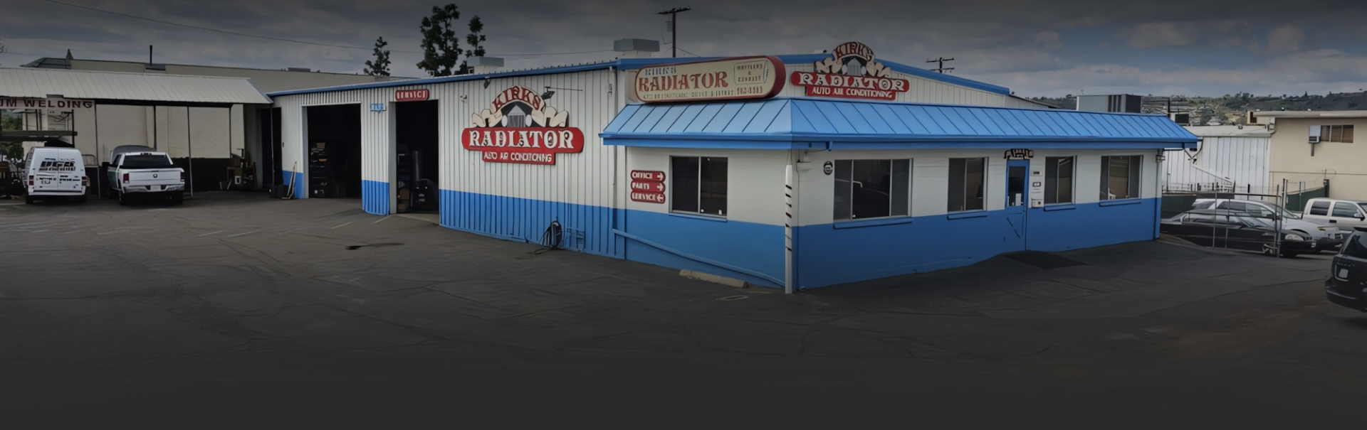 A white and blue building with a blue roof and a lot of cars parked in front of it. | Kirk's Radiator & Auto A/C Inc.