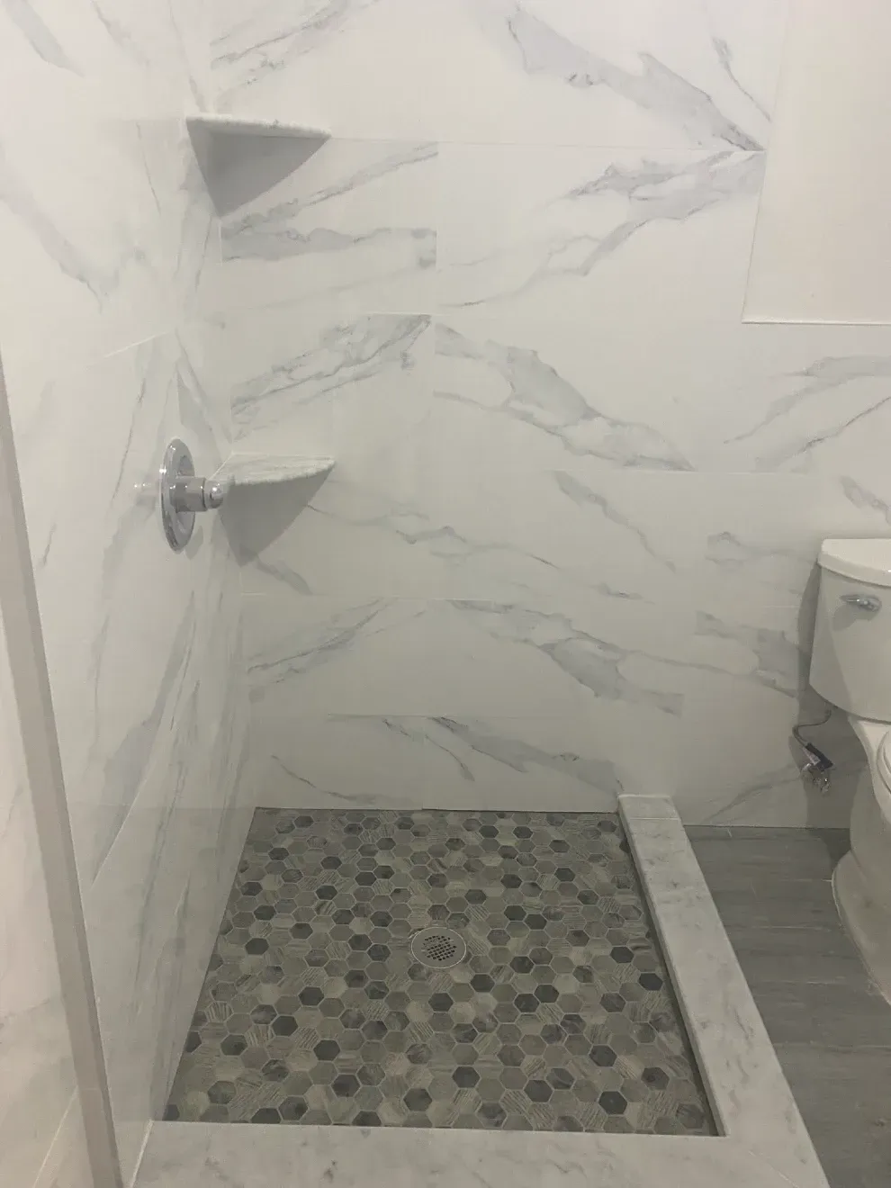 Shower stall with marble-like tile walls, two corner shelves, and mosaic tile floor. A toilet is visible on the right.