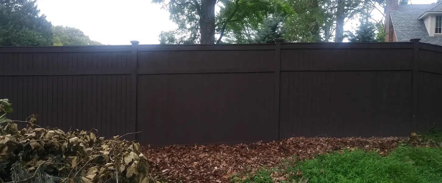 Brown wooden fence in a yard with trees and house in background.