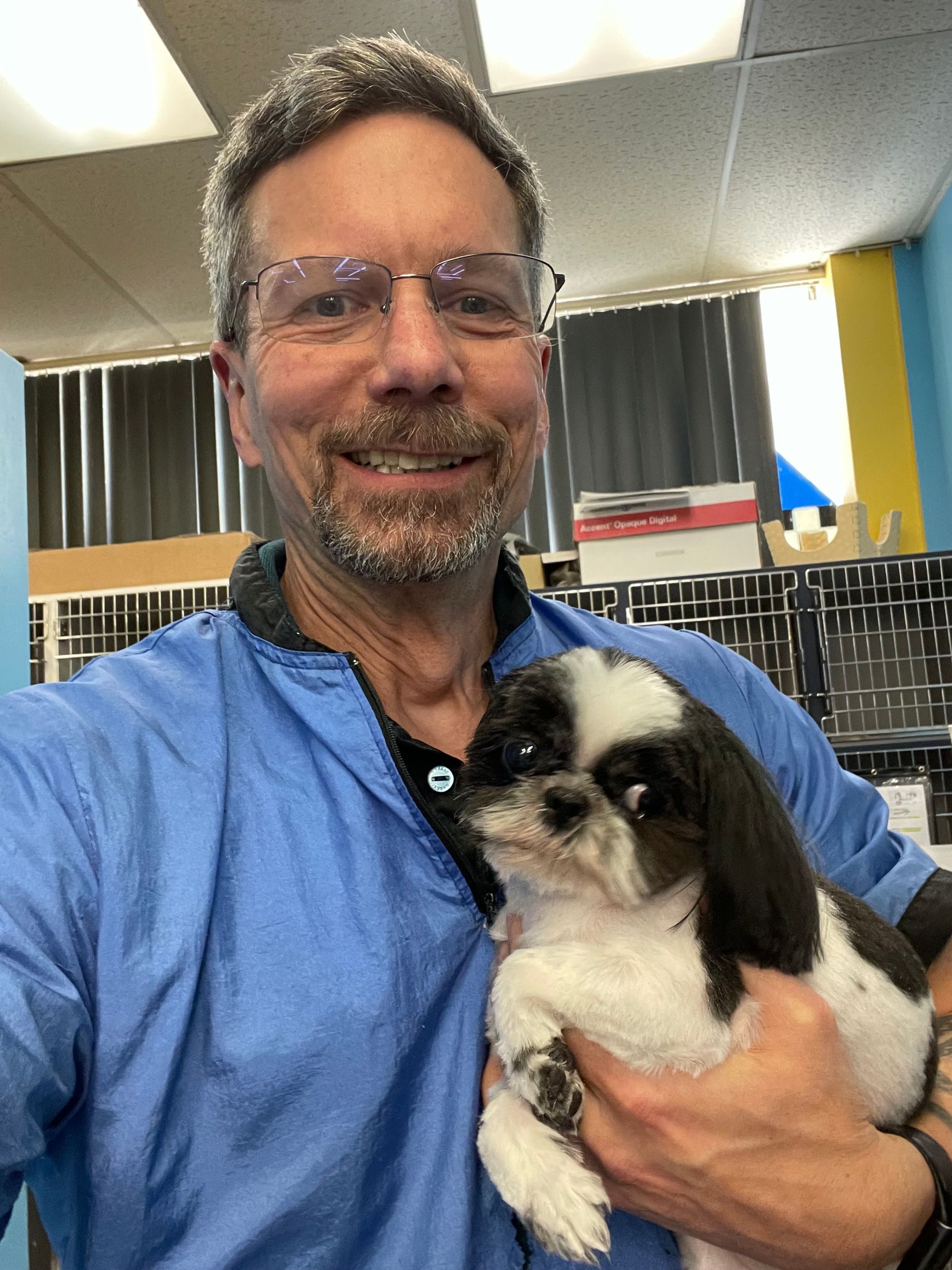 Man holding a black and white Shih Tzu. Man is smiling with glasses on. Indoors.
