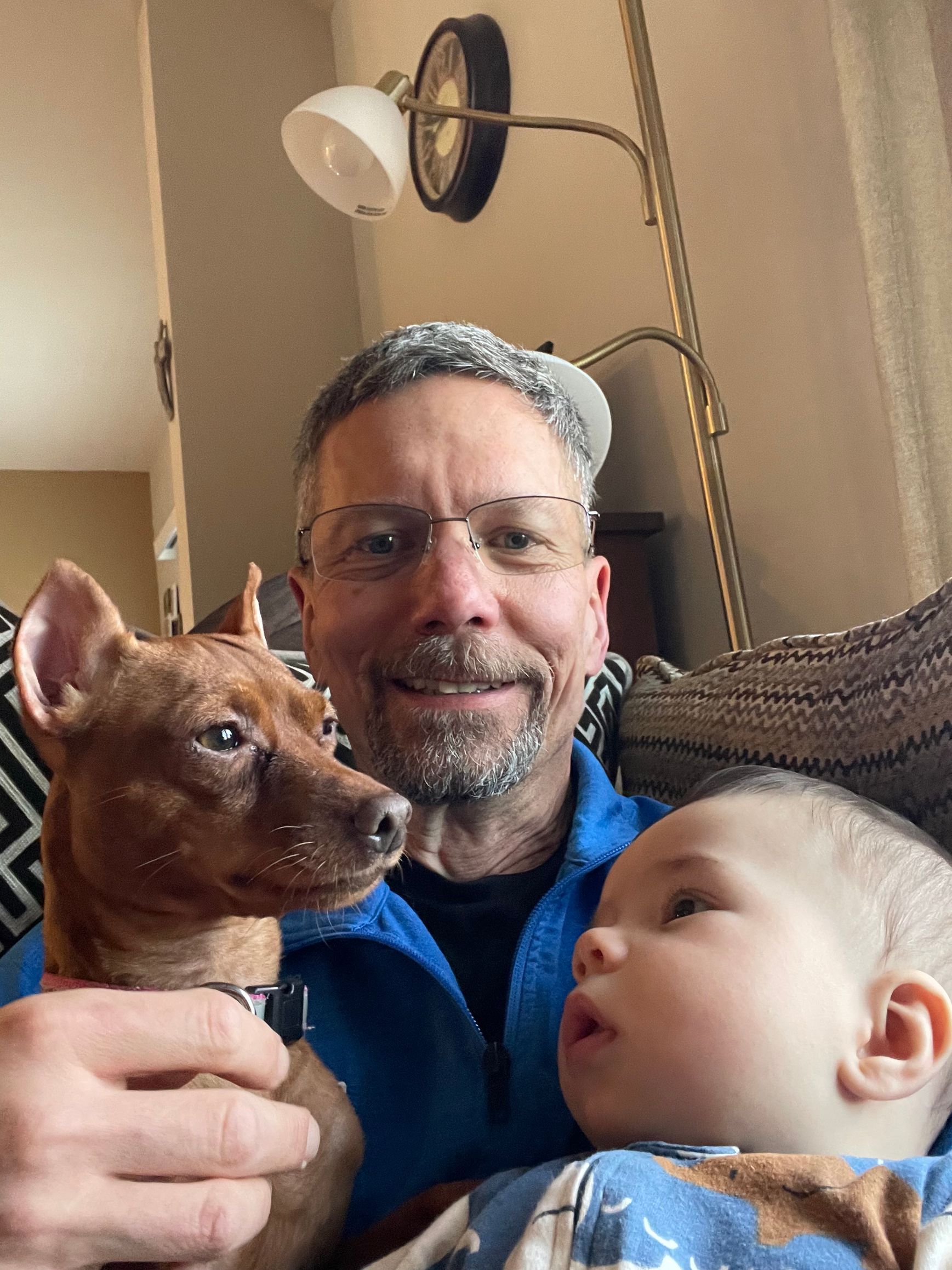 A man is holding a baby and a dog while sitting on a couch.