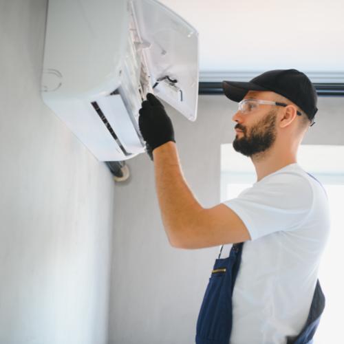HVAC technician in black cap and gloves repairs an AC unit on a wall.
