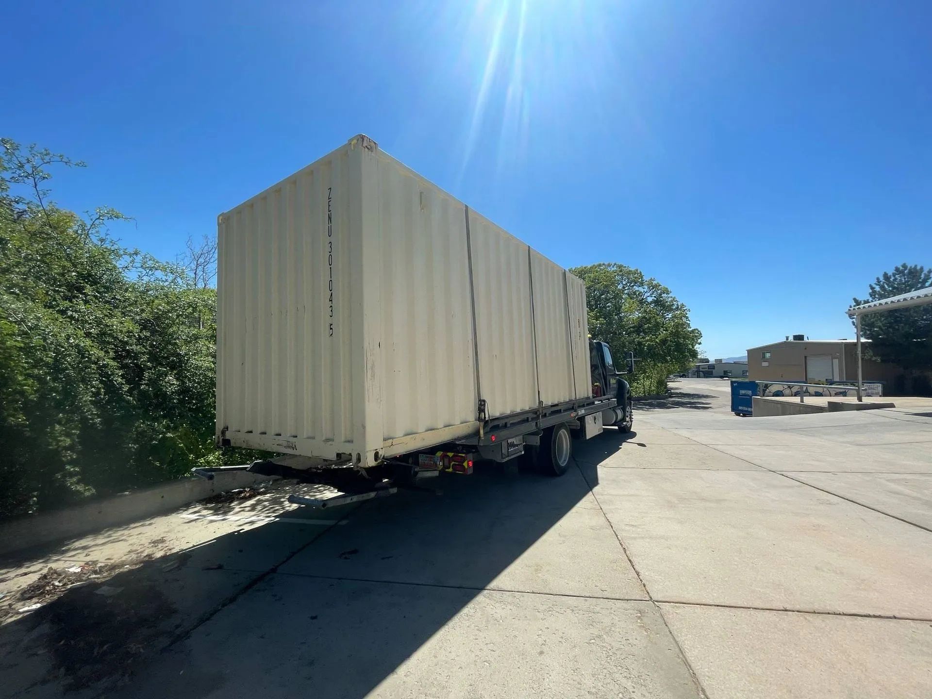 A truck with a large, beige shipping container on a paved surface under a bright blue sky.